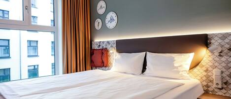 A hotel room with a bed, curtains, wall clocks, and a nightstand with a remote control.