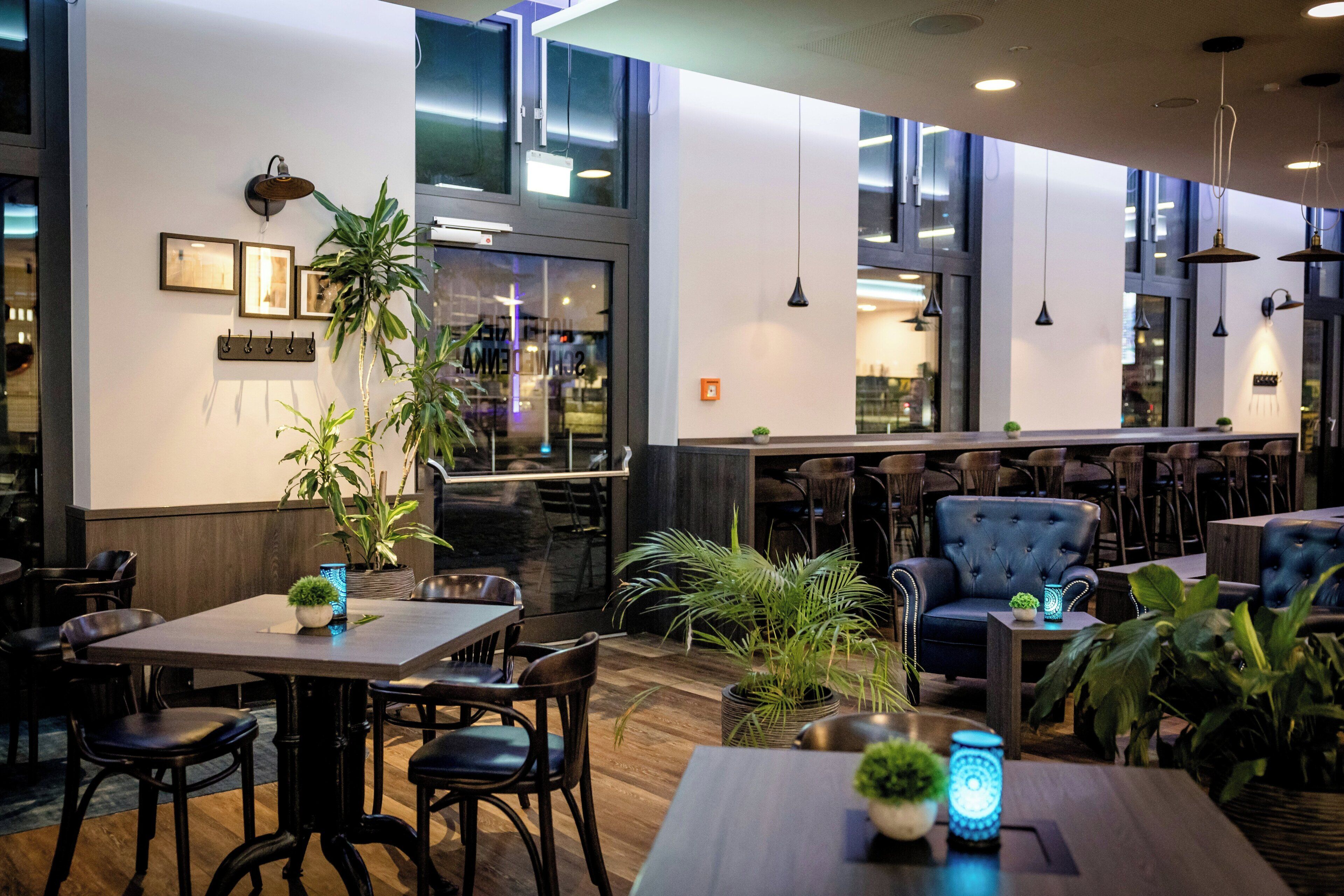 A modern restaurant interior with wooden tables, chairs, potted plants, and pendant lighting.