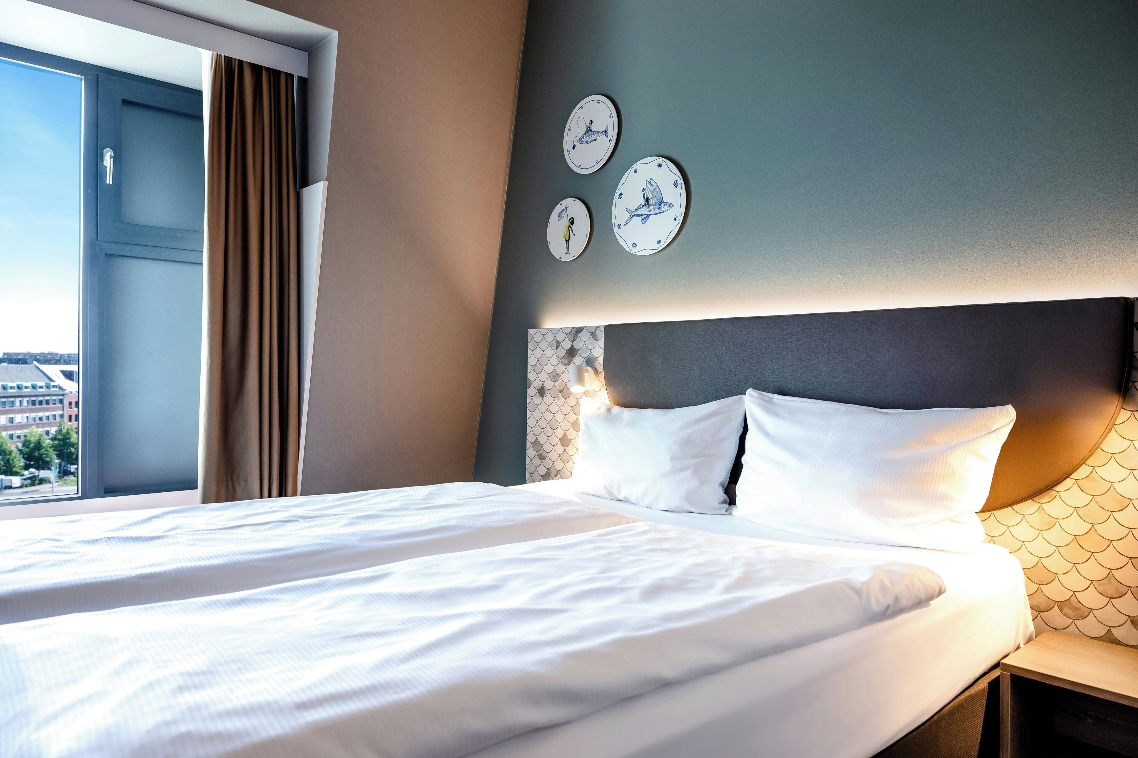 A hotel room with a bed, a nightstand, a window with curtains, and wall decor.