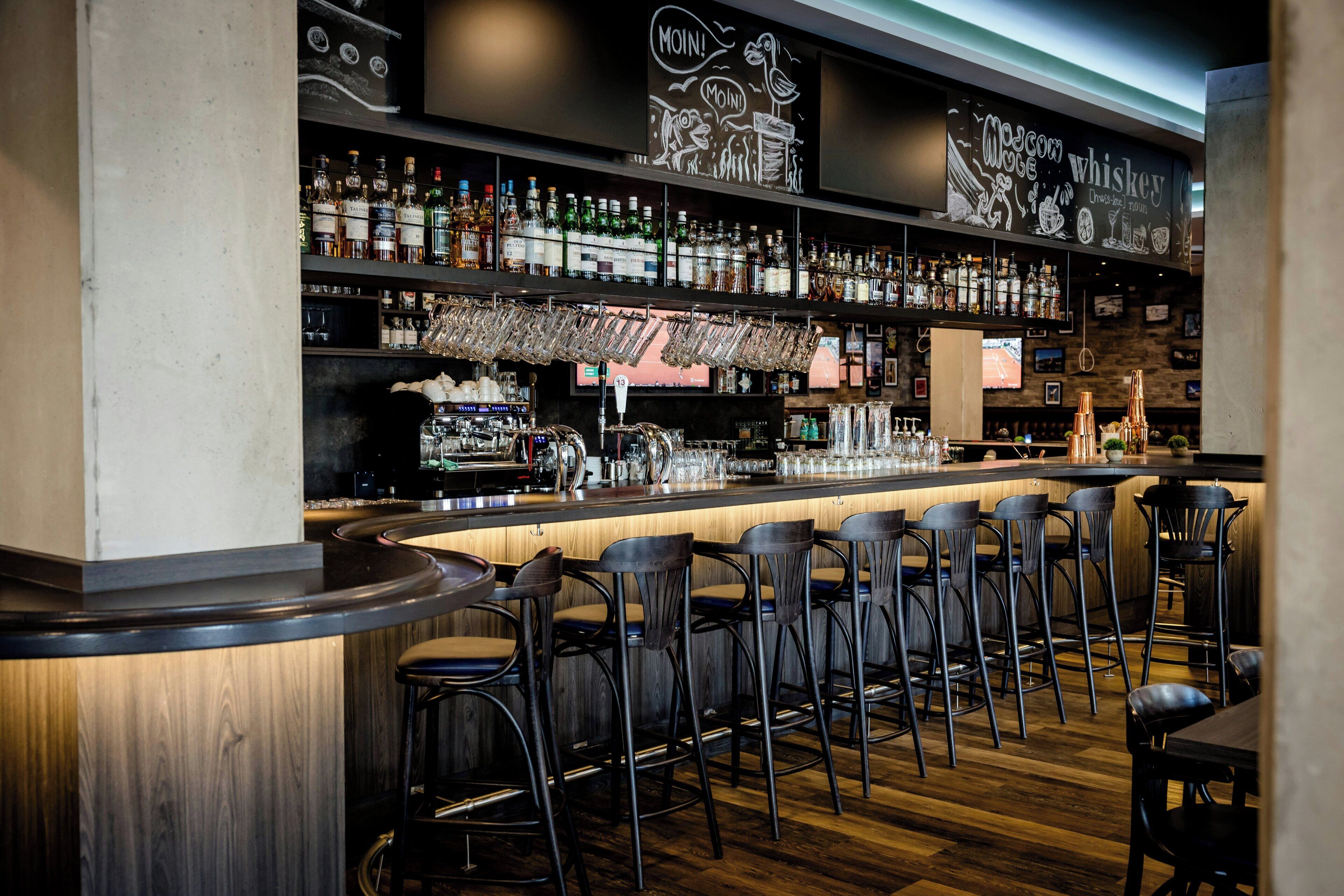 Zona de bar con varios taburetes, estantes con botellas y una barra.