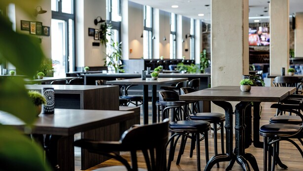 A modern restaurant interior with high ceilings, large windows, and a mix of wooden and metal furniture. - Hotel Kiel-Schwedenkai (Kiel)