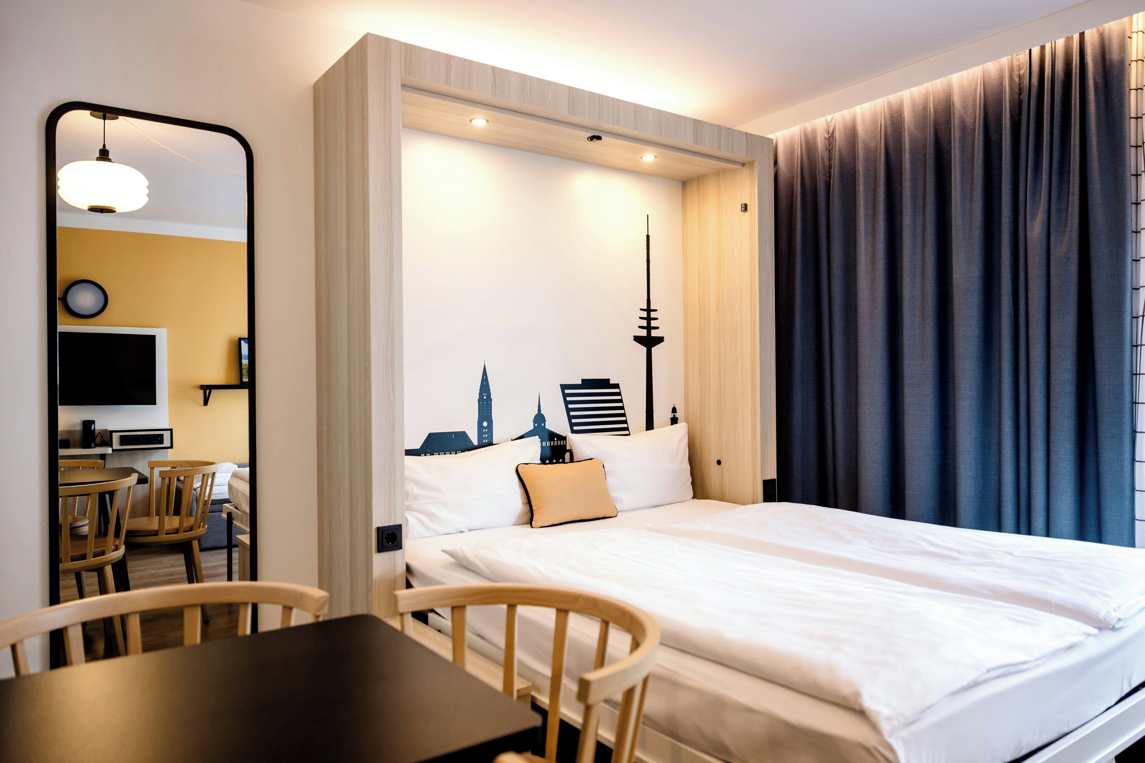 A hotel room with a bed, a desk, chairs, a mirror, and a wall-mounted TV.
