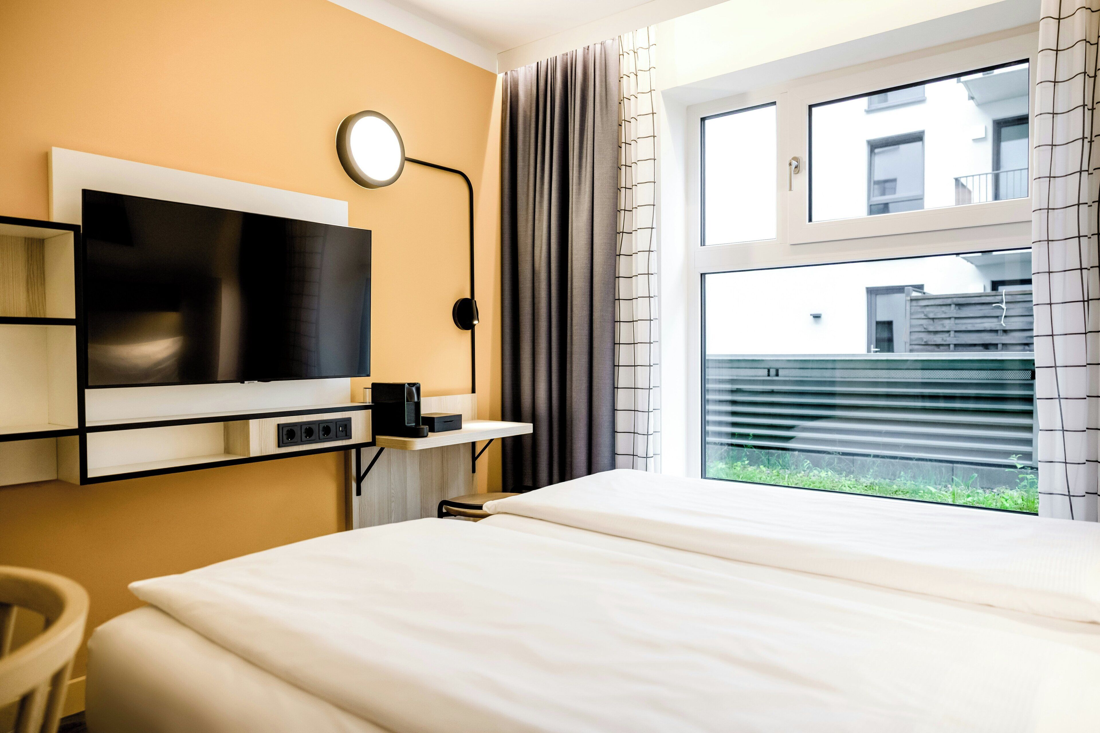 Habitación de hotel con cama, televisión montada en la pared, escritorio con lámpara y ventana con cortinas.