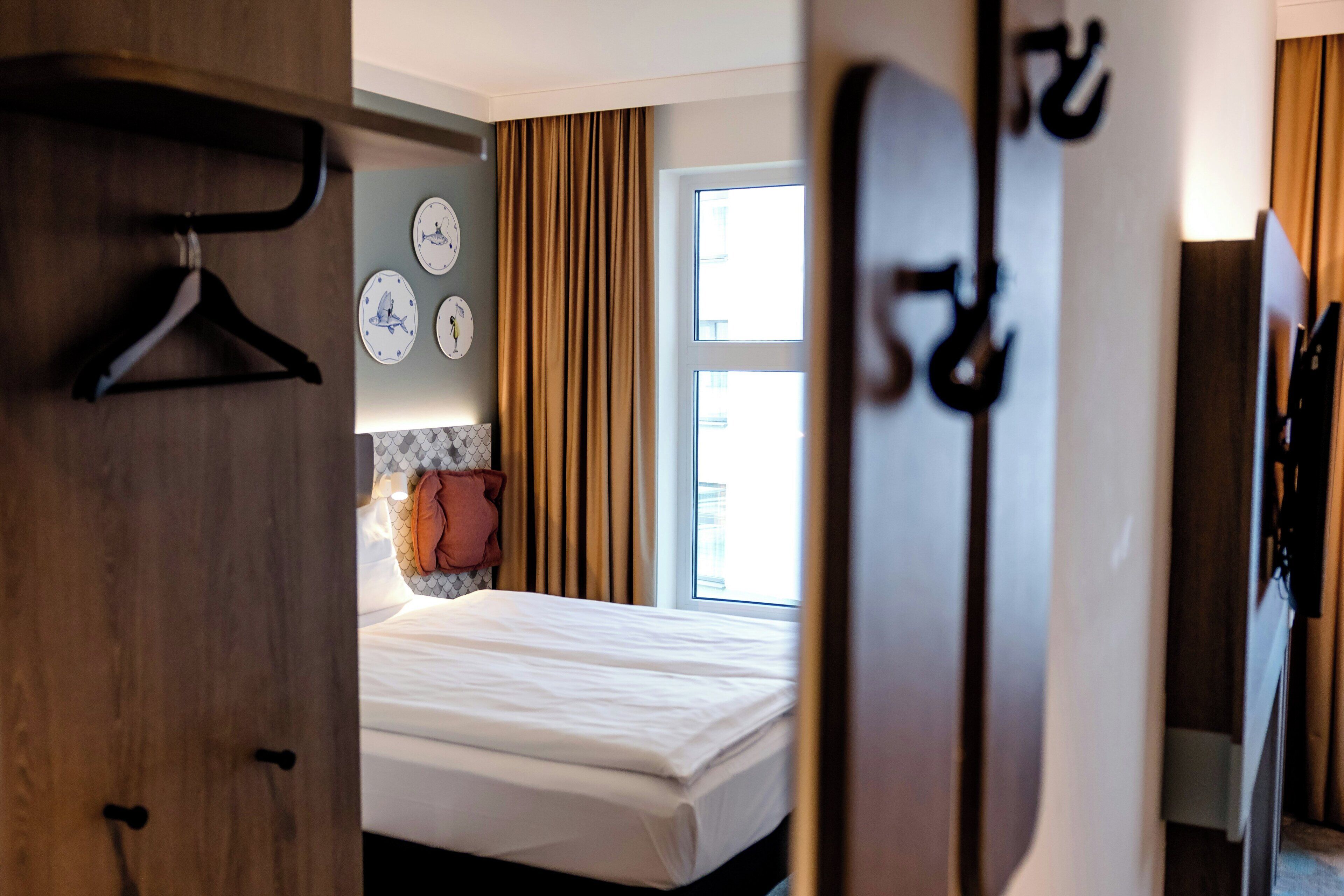 Una habitación de hotel con cama, armario, espejo y ventana con cortinas.
