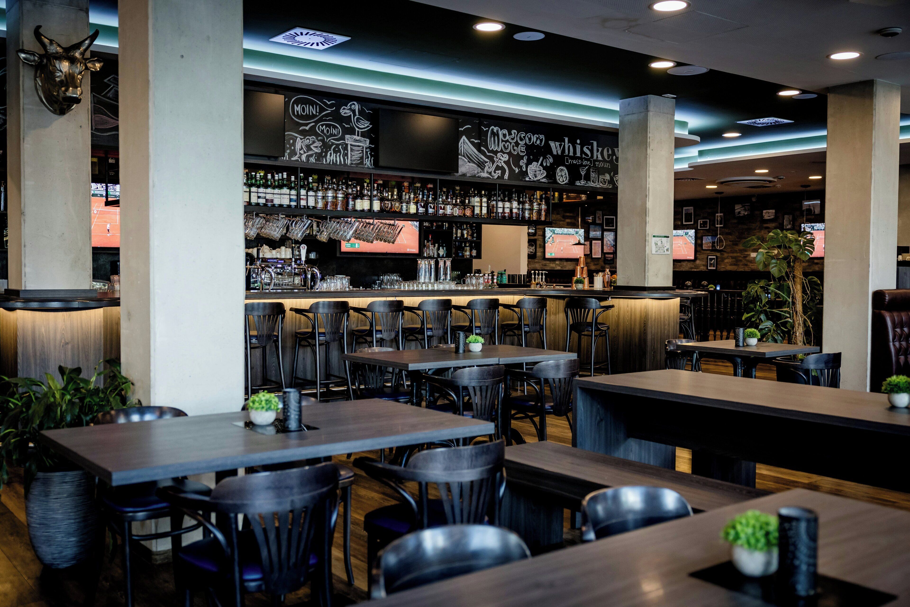 A bar area with multiple tables and chairs, a bar counter with various bottles, and a television screen.