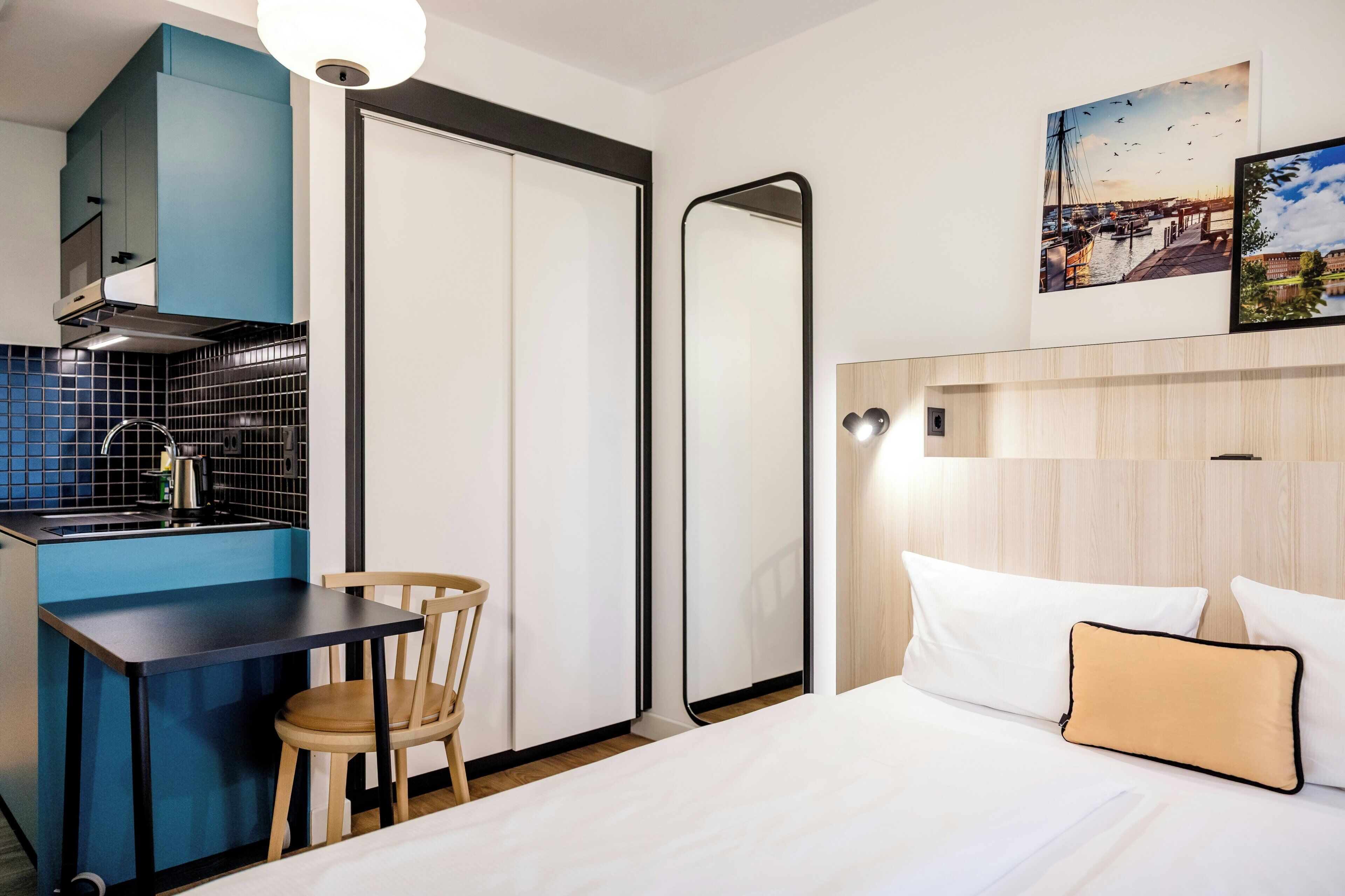 Habitación de hotel compacta con kitchenette, una cama con ropa blanca, una silla de madera y un espejo.