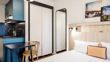 Habitación de hotel compacta con kitchenette, una cama con ropa blanca, una silla de madera y un espejo.