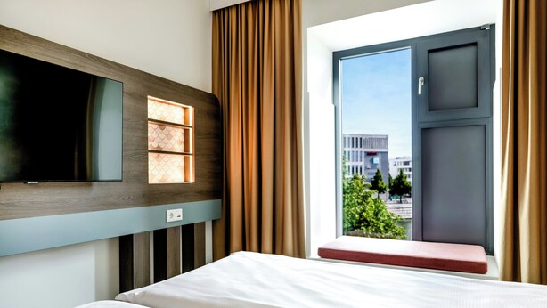 A hotel room with a bed, a television mounted on the wall, a window with curtains, and a view of the outside. - Hotel Kiel-Schwedenkai (Kiel)