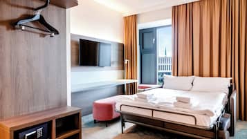 Ein Hotelzimmer mit einem Bett, einem an der Wand befestigten Flachbildfernseher, einem Holztisch ebenfalls mit Flachbildfernseher und einem Fenster mit Vorhängen.