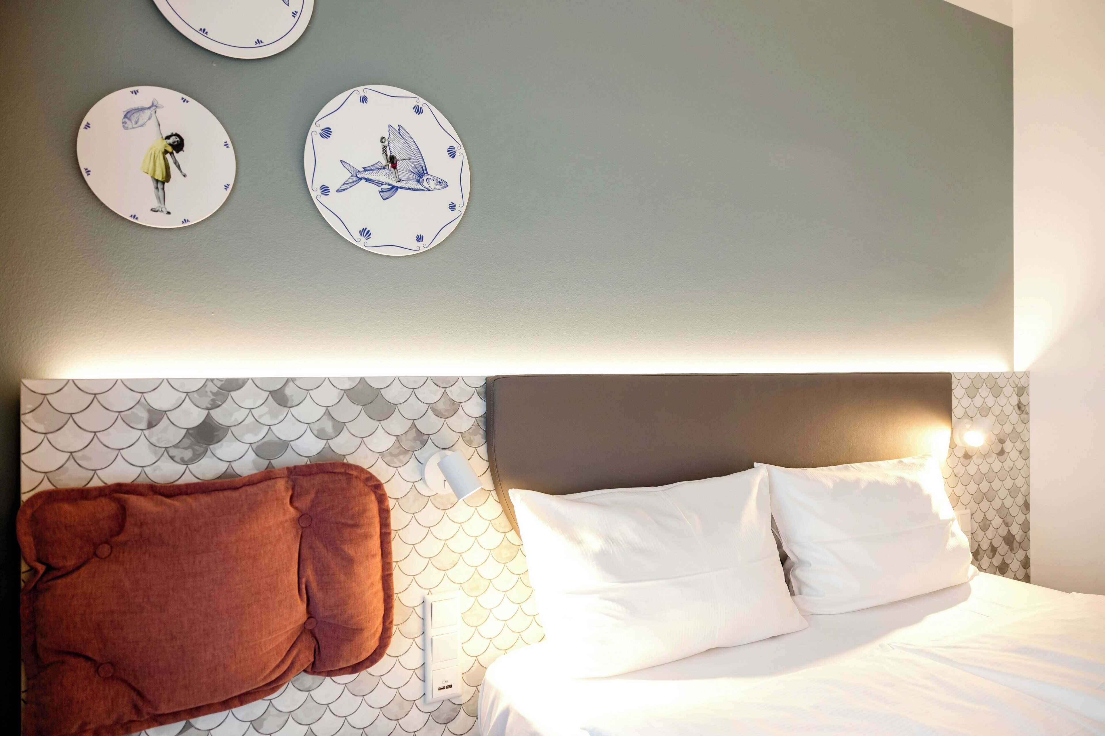 A hotel room with a bed, white pillows, a brown blanket, and wall clocks with fish illustrations.