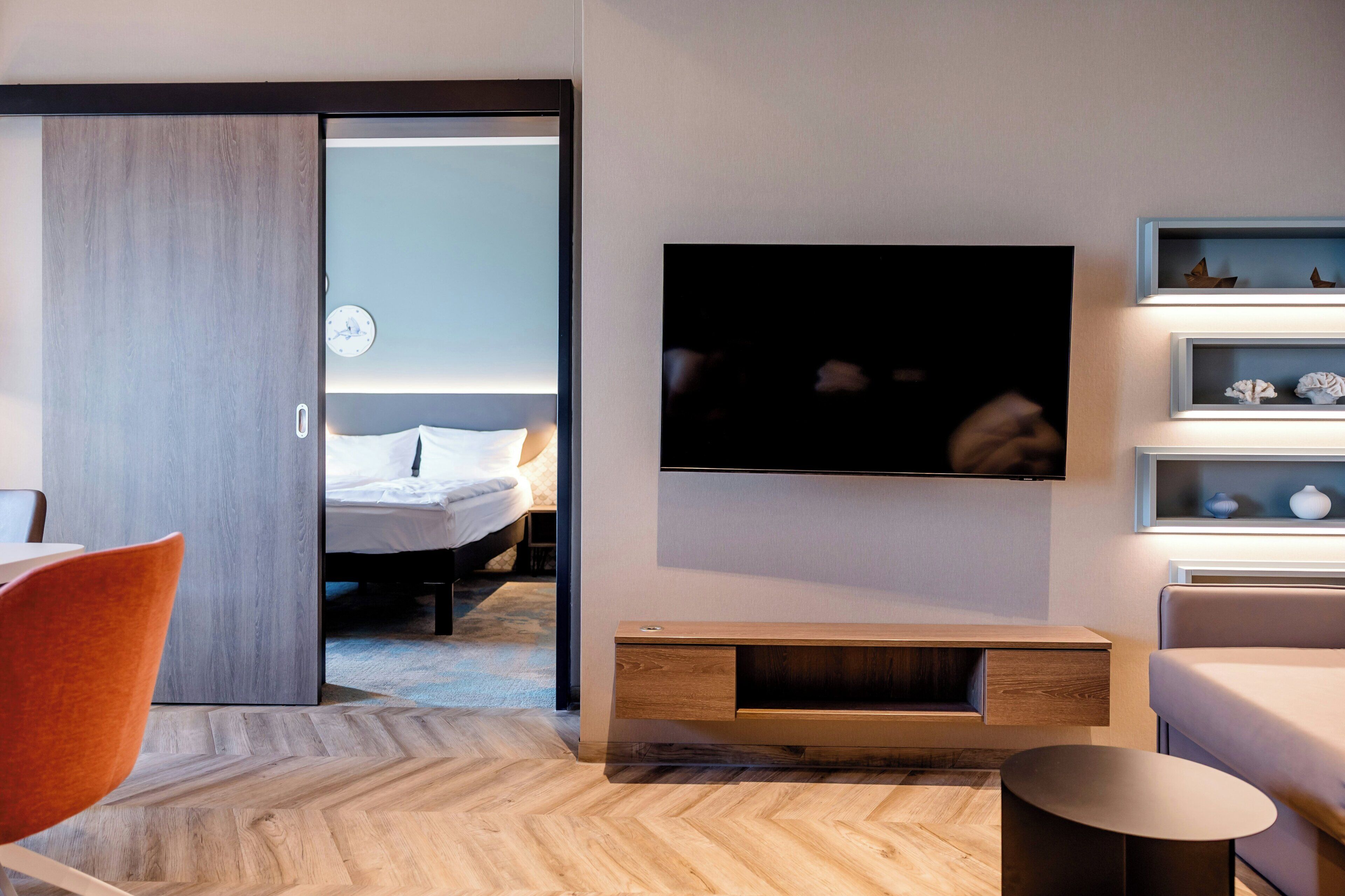 A modern hotel room with a flat-screen TV mounted on the wall, a wooden floor, a bed with white bedding, and a seating area with a chair and a small table.