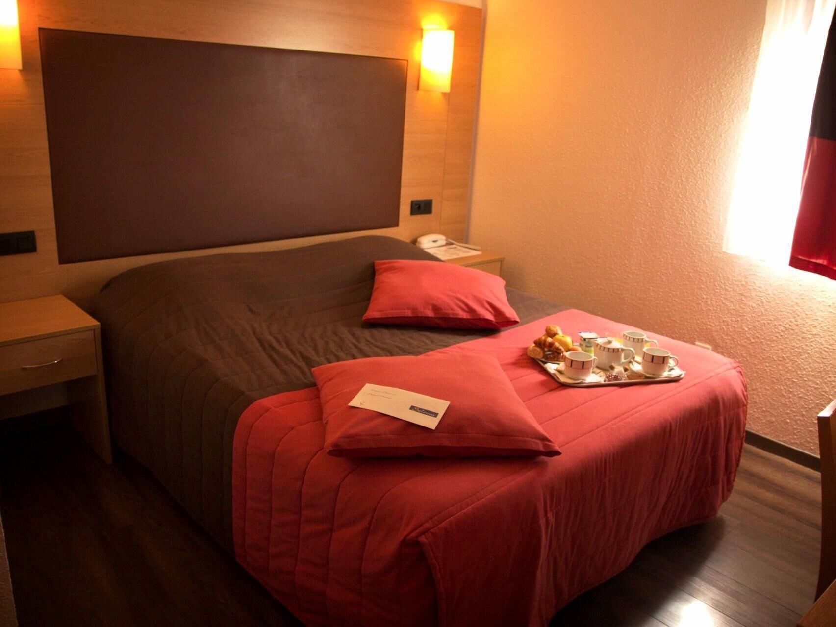 Een hotelkamer met een groot bed, een nachtkastje, een dienblad met ontbijtartikelen en een rood kussen.
