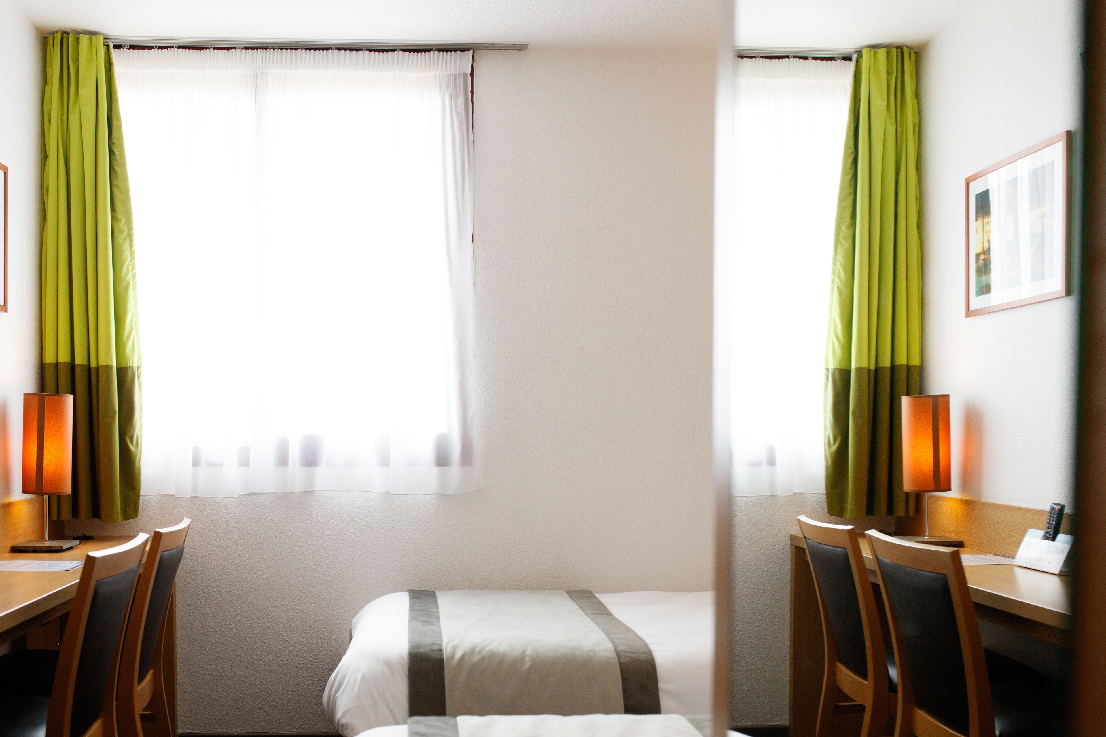 Une chambre d’hôtel avec un lit, deux chaises, un bureau, une lampe et une fenêtre avec des rideaux.