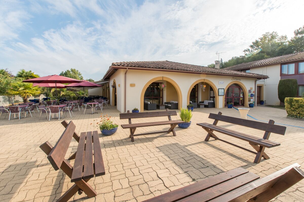 An outdoor patio area with wooden benches, a covered seating area with red umbrellas, and a building with arched doorways.