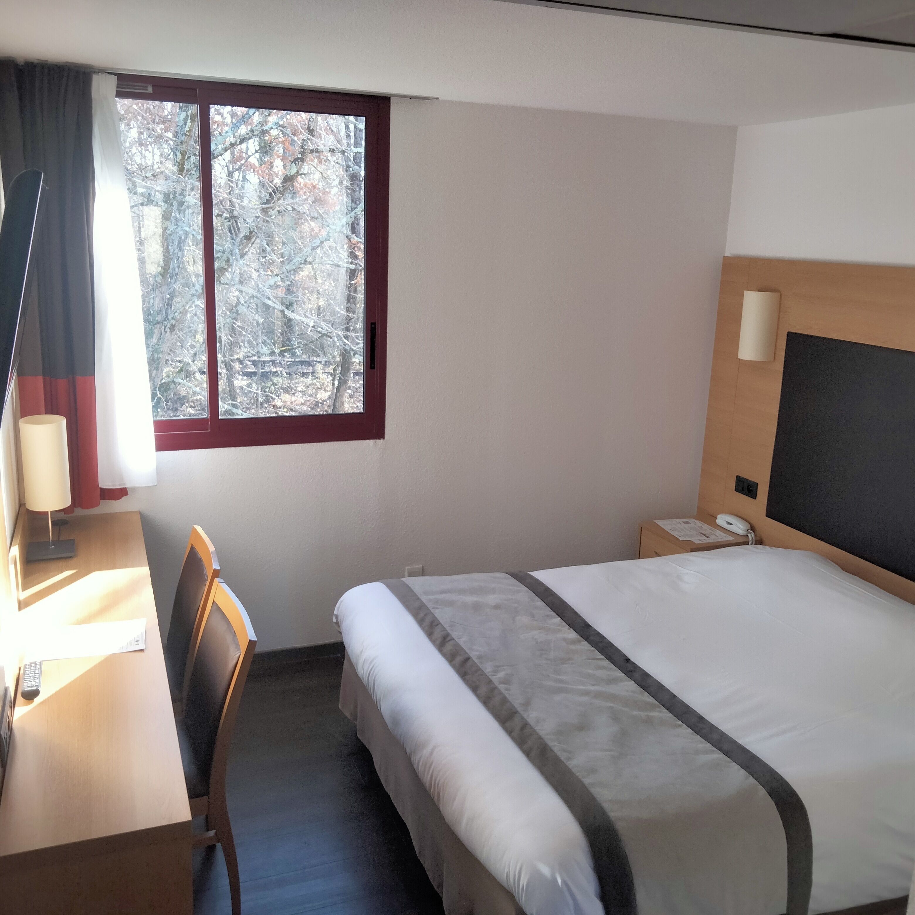 A hotel room with a bed, a desk, a chair, a lamp, a telephone, and a window with a view of trees.