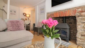 A cozy living room with a fireplace, a sofa, a TV, and a vase of pink roses. - Nookie Cottage (Dartmouth)