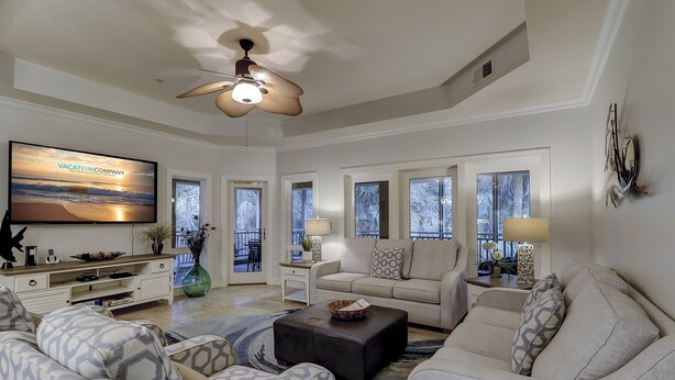 A spacious living room with a ceiling fan, a flat-screen TV, and a large window with a view of trees. - 8126 Wendover Dunes - Palmetto Dunes Oceanside Community (Hilton Head Island)