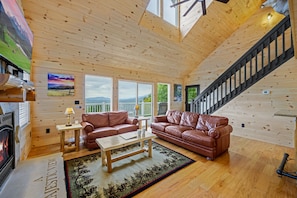 A cozy cabin interior with wooden walls, a fireplace, leather sofas, a dining table, and a staircase.