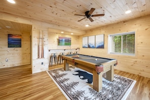A wooden interior with a pool table, bar stools, and a ceiling fan.