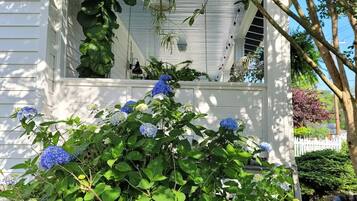A white house with a covered porch, surrounded by lush greenery and hydrangea bushes.