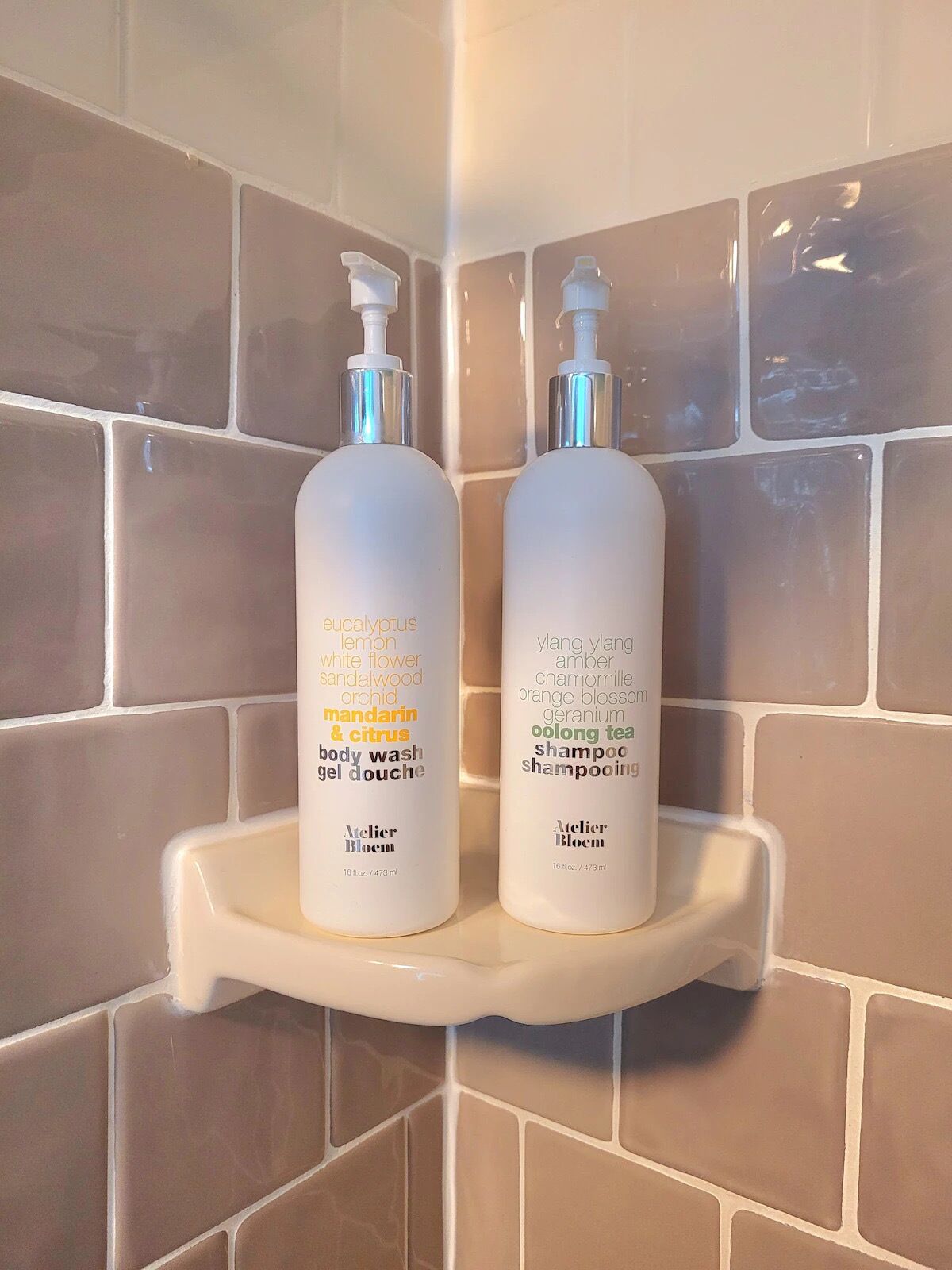 Two bottles of Atelier Bloom body wash and shampoo on a white shelf against a tiled wall.
