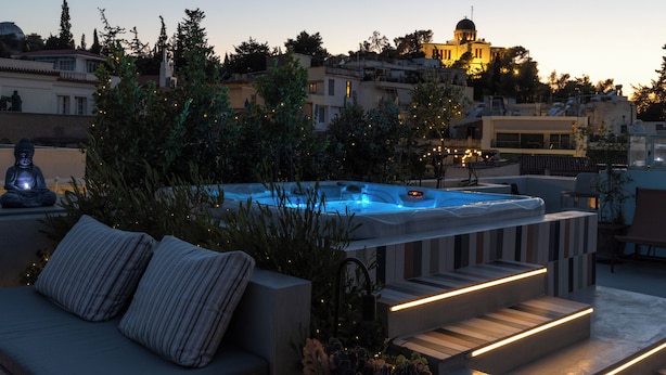 En tagterrasse med jacuzzi, udendørs møbler og udsigt over bybilledet i skumringen.