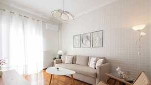 En moderne stue med en sofa, et glasbord, trægulv, en hvid væg med kunst og en loftlampe.