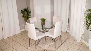 A dining area with a glass table surrounded by four white chairs, a potted plant, and white curtains.