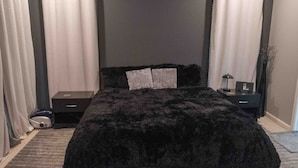 A bedroom with a black bedspread, two pillows, a bedside table, a lamp, and a vacuum cleaner.
