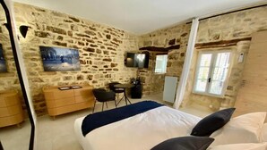 A room with a stone wall, a bed with pillows, a desk with a chair, a TV, and a window with curtains. - Grand Luxe SENSATION Suite with private Jacuzzi (Martignargues)