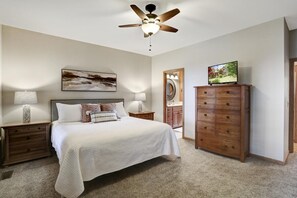 Une chambre à coucher comprenant un lit, des tables de chevet, une commode, un ventilateur de plafond, un miroir fixé au mur et un téléviseur.