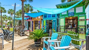 A wooden deck with blue Adirondack chairs, a potted plant, and a storefront with a 'Pay Here' sign. - Sea You Soon 1 Bedroom Condo by RedAwning (Santa Rosa Beach)
