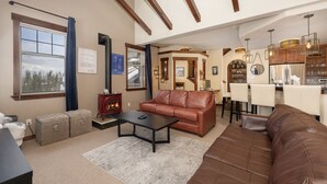 A cozy living room with a fireplace, leather sofas, a coffee table, and a dining area with a kitchen in the background. - Cozens Pointe B-301 3 Bedroom Condo by RedAwning (Fraser)