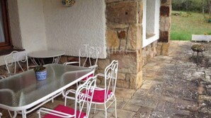 Une terrasse couverte avec des murs en pierre, une table avec des chaises et un petit jardin visible à travers la fenêtre.