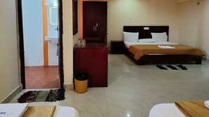 A hotel room with a bed, a desk, a trash bin, and a door leading to a bathroom. - Hotel Geetha International (Thoothukudi)