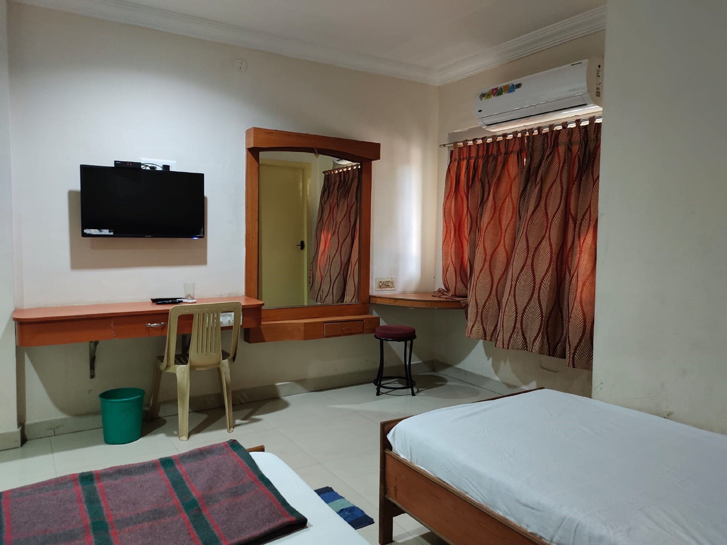Un dormitorio con cama, escritorio y silla, televisor, ventana con cortinas y aire acondicionado.