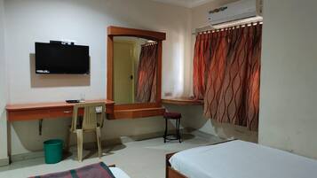 Un dormitorio con cama, escritorio y silla, televisor, ventana con cortinas y aire acondicionado.