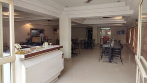 A spacious dining area with a long table and chairs, a reception desk with a computer and decorative items, and a ceiling fan. - Hotel Geetha International (Thoothukudi)
