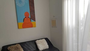 Una sala de estar con un sofá negro, dos almohadas blancas, una mesita pequeña con un control remoto y un cuadro en la pared.