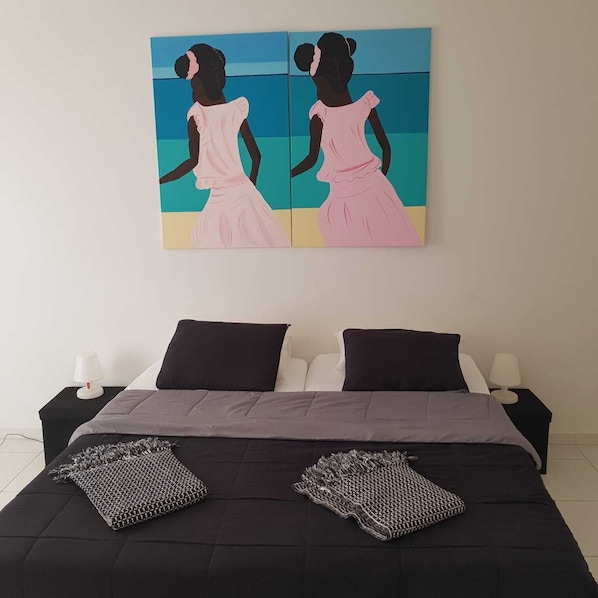 A bedroom with a bed, two black nightstands, two white lamps, and a painting of two women on the wall.