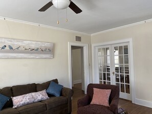 A living room with a brown sofa, a patterned armchair, a ceiling fan, and a door leading to another room.