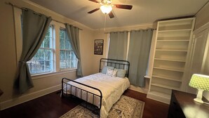 A bedroom with a bed, a ceiling fan, a window with curtains, and a built-in shelving unit. - Beautiful 1930s brick home in historic district. (Sumter)