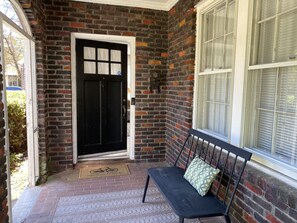 A brick exterior with a black door, a bench with a cushion, and a window with blinds. - Beautiful 1930s brick home in historic district. (Sumter)