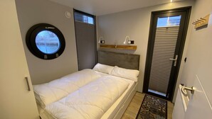 Ein Schlafzimmer mit einem Bett, einem runden Fenster, einer Tür mit Griff und einem kleinen Regal mit einer Lampe.