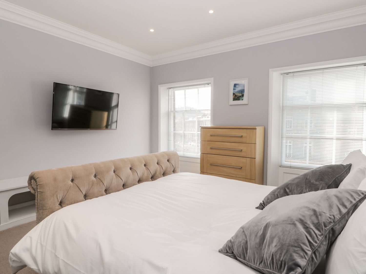 A bedroom with a large bed, a wooden dresser, a TV mounted on the wall, and two windows with blinds.