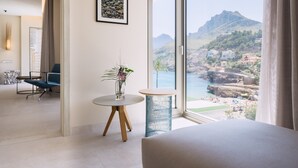 Habitación de hotel con un ventanal que ofrece vistas a un paisaje costero, una cama, una pequeña mesa redonda con un florero y una silla.