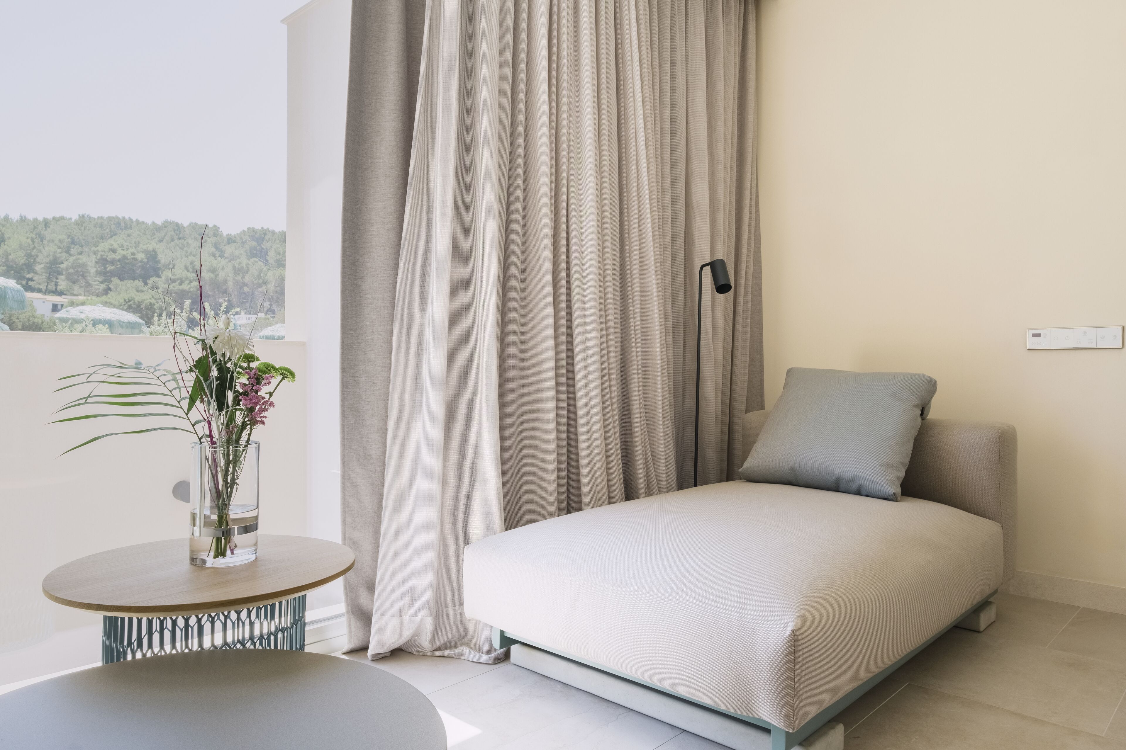 Un dormitorio moderno con una cama, una mesita redonda con un jarrón de flores y vistas al exterior con vegetación.