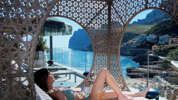Una mujer descansando en una silla lounge con una copa de vino, con vista a la costa, una piscina y montañas al fondo.