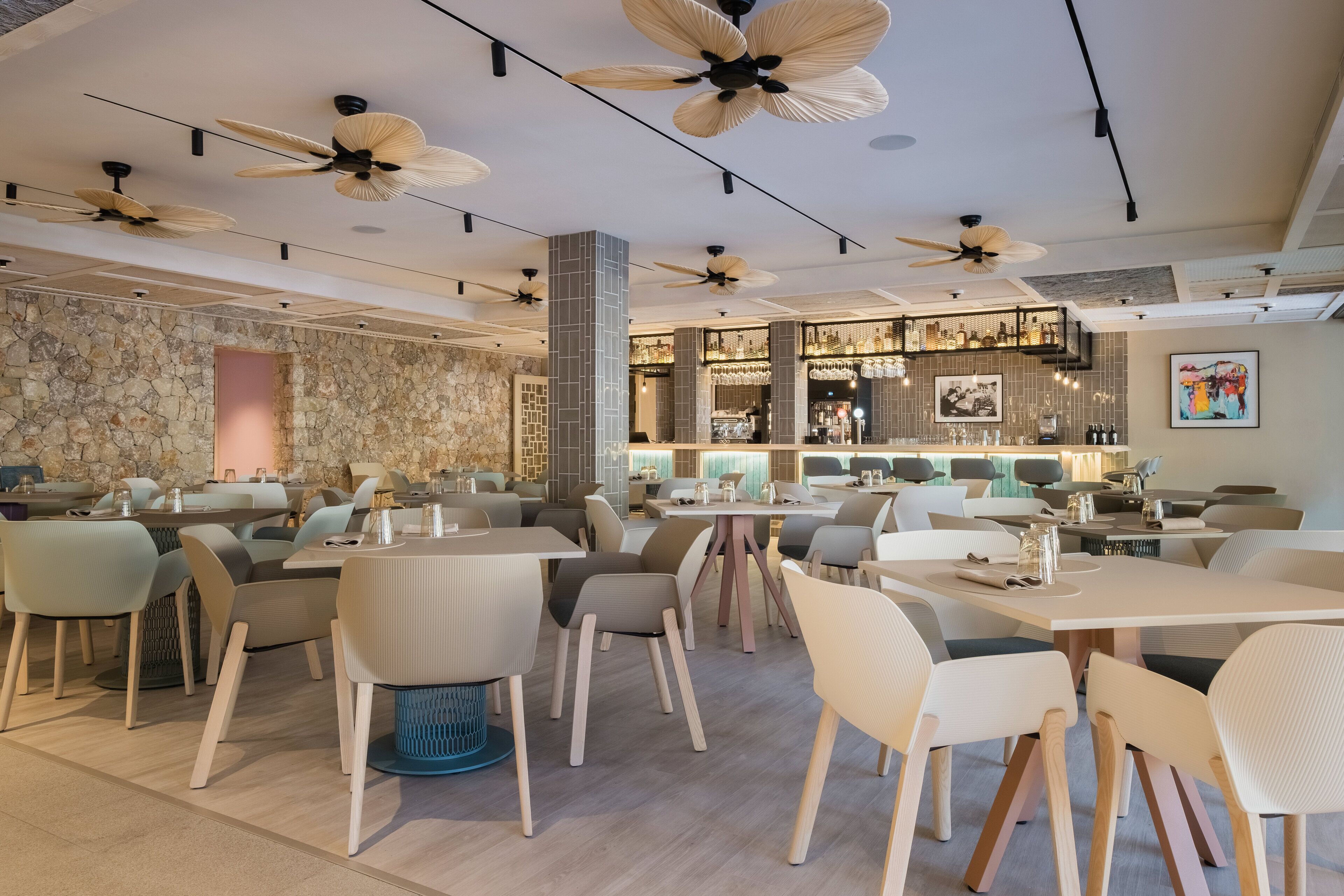Een modern restaurant met meerdere eettafels en stoelen, plafondventilatoren en een bar met flessen op planken.
