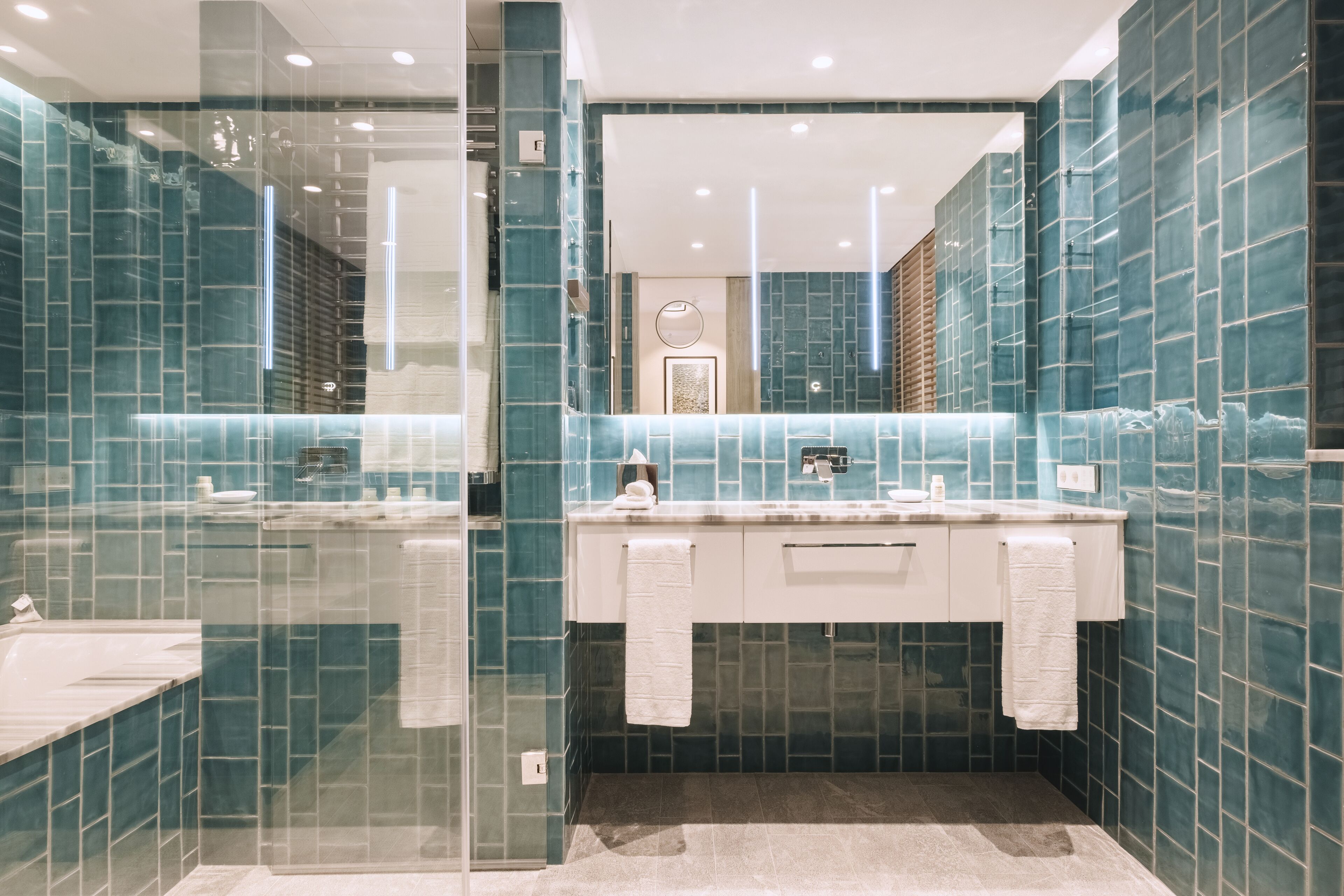 Un baño moderno con cabina de ducha de vidrio, tocador blanco con espejo y paredes con azulejos color turquesa.
